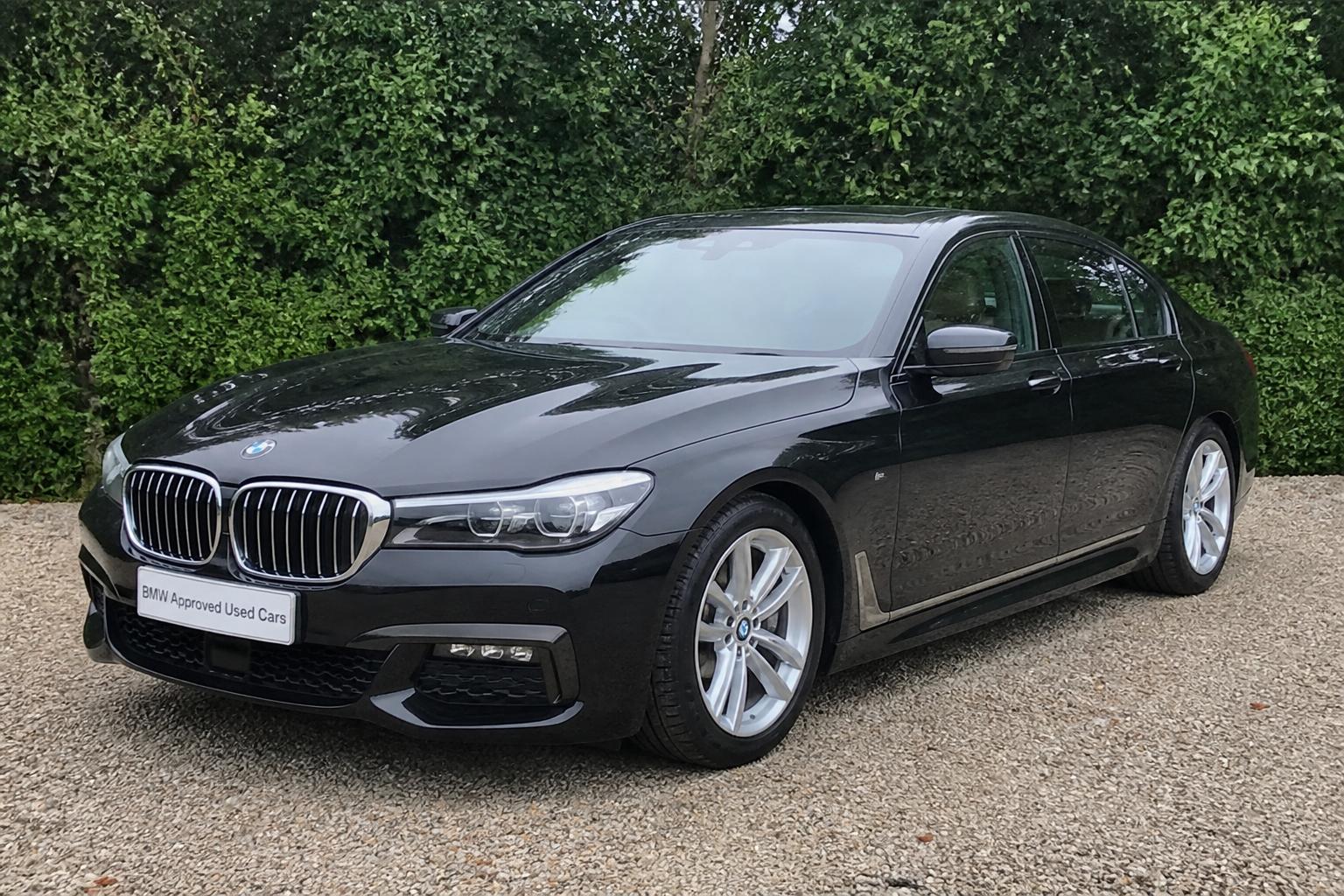 Black BMW 7 Series parked on gravel outdoors with a bush behind it